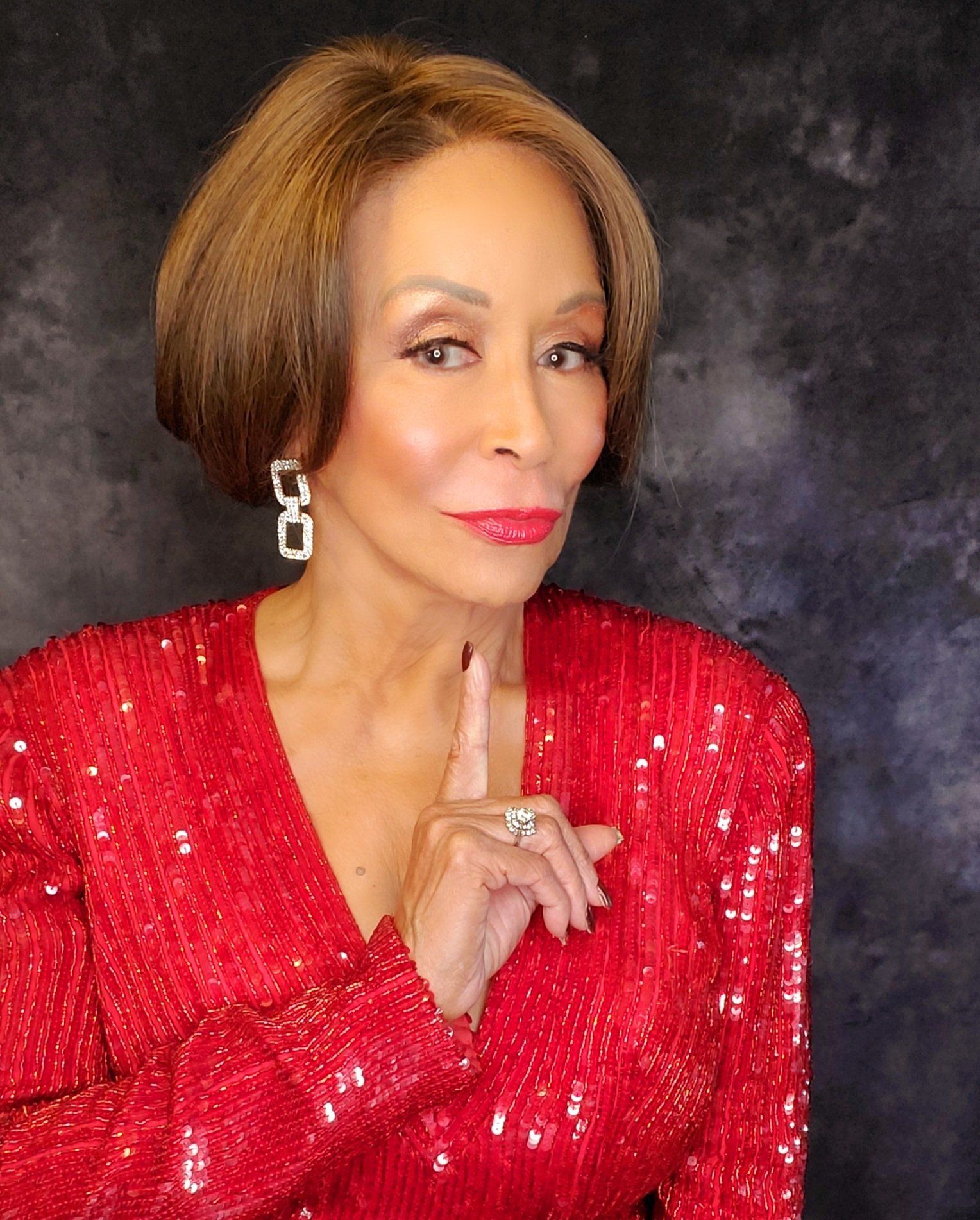 Portrait of a woman in a red sequined dress against a dark backdrop, smiling gently with hand near chin