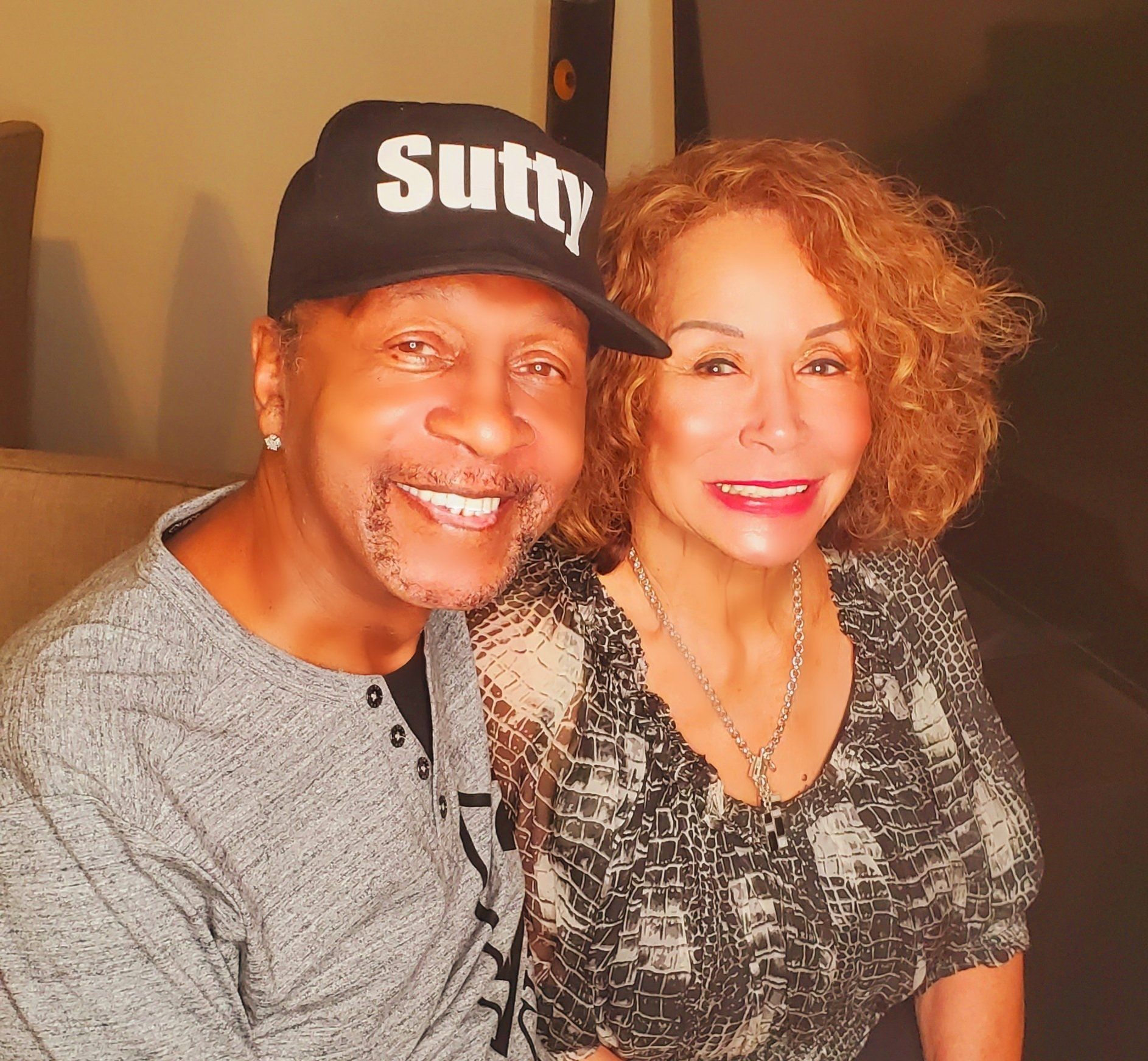 Two smiling people seated on a couch, wearing casual clothes, with warm indoor lighting.