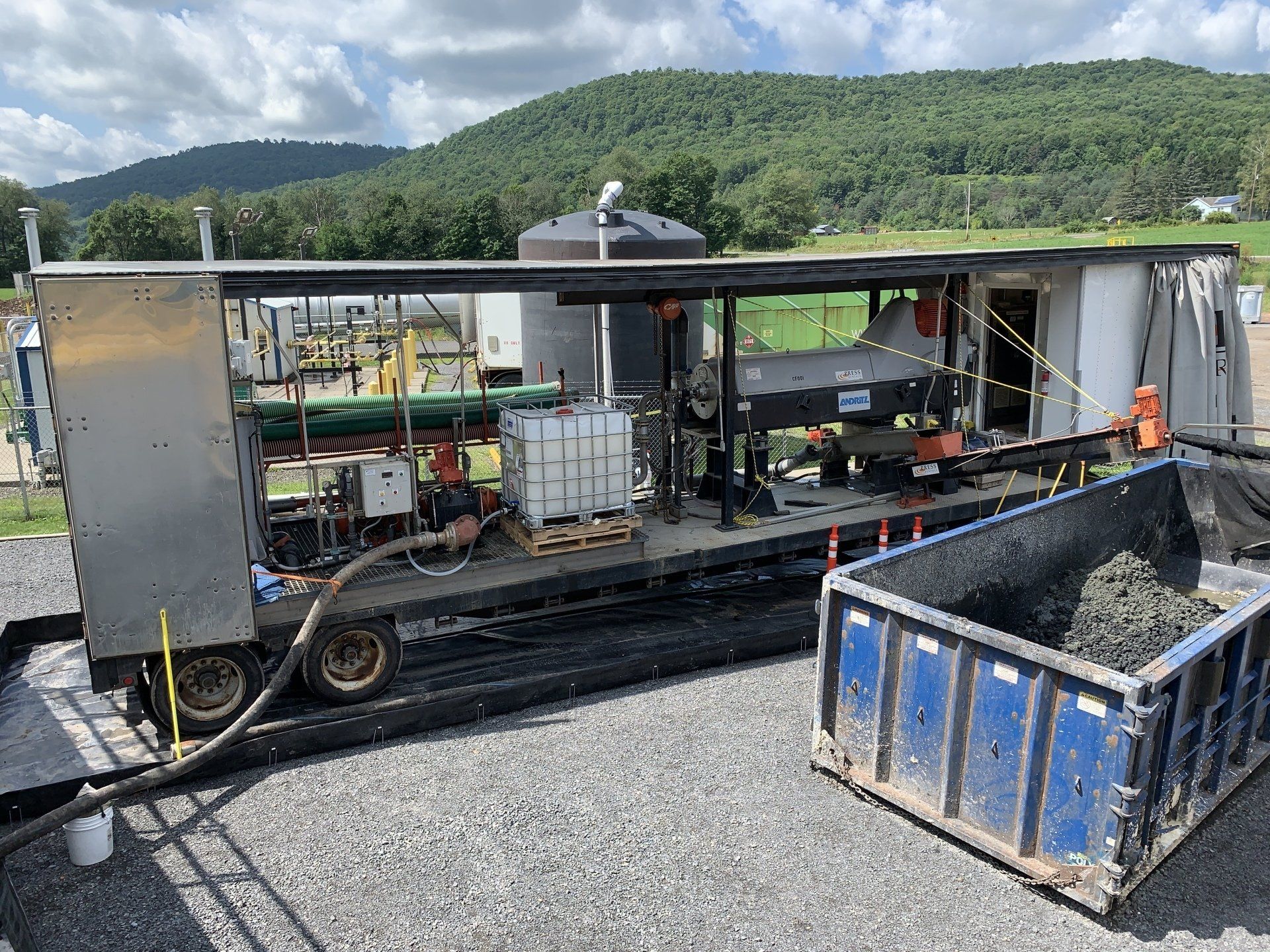 A trailer is sitting on top of a gravel road next to a dumpster.
