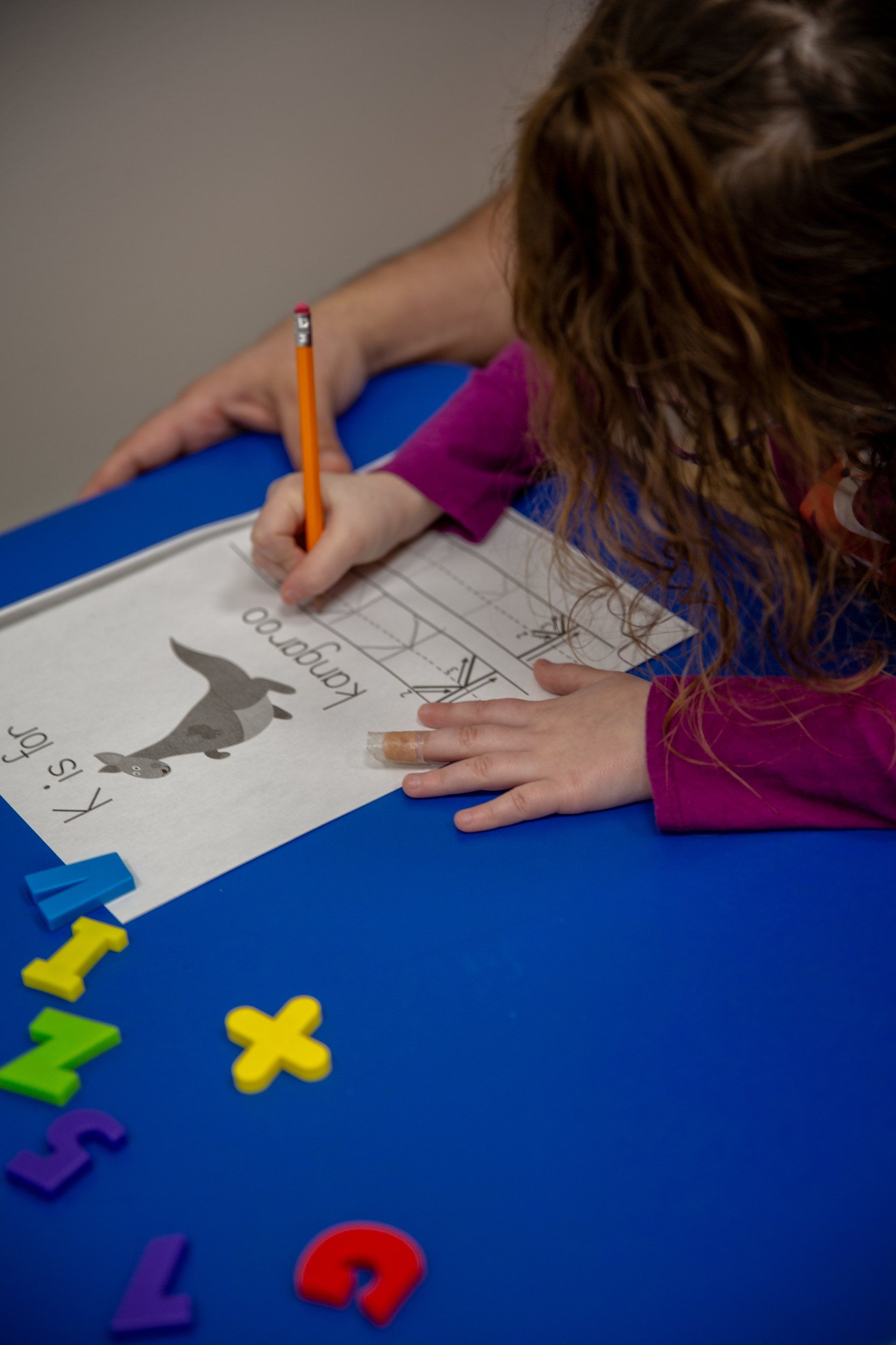 Child writing in a workbook at a blue table with colorful letter and number tiles nearby