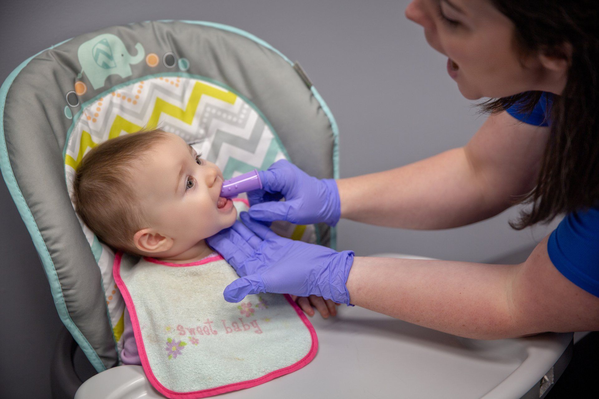 Caregiver feeding a baby in a high chair with a spoon, wearing purple gloves