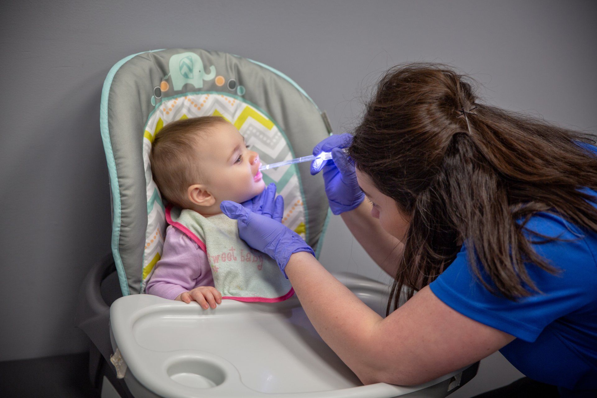 Caregiver in blue gloves examines a baby in a high chair with a penlight