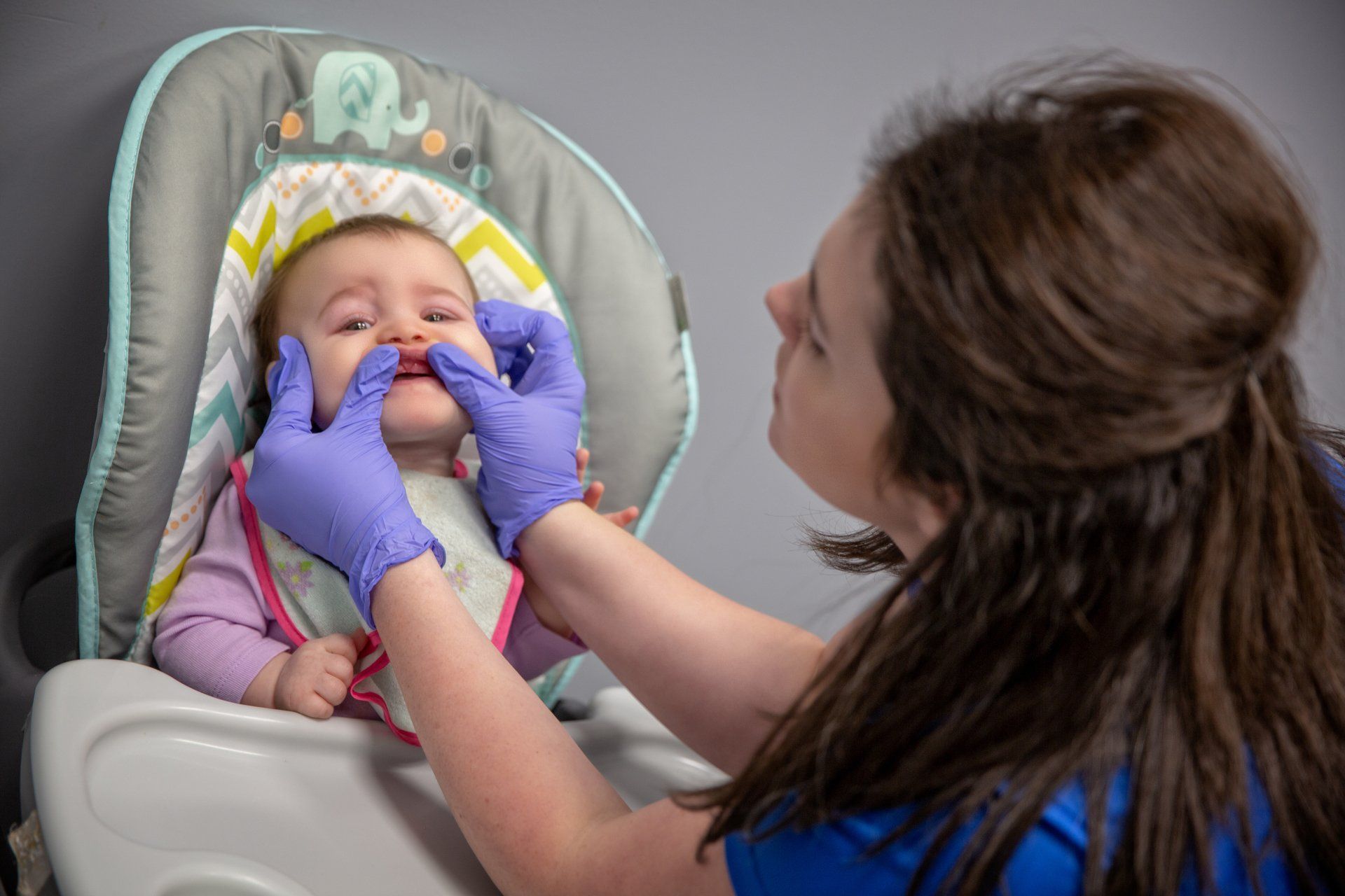 Nurse examining a baby in a high chair, wearing purple gloves and adjusting the infant’s mouth.