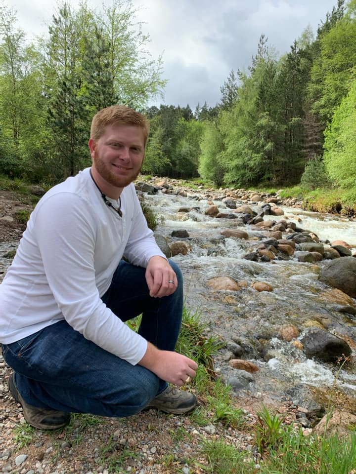 Person crouching beside a rocky stream in a green forest, wearing a white shirt and jeans.