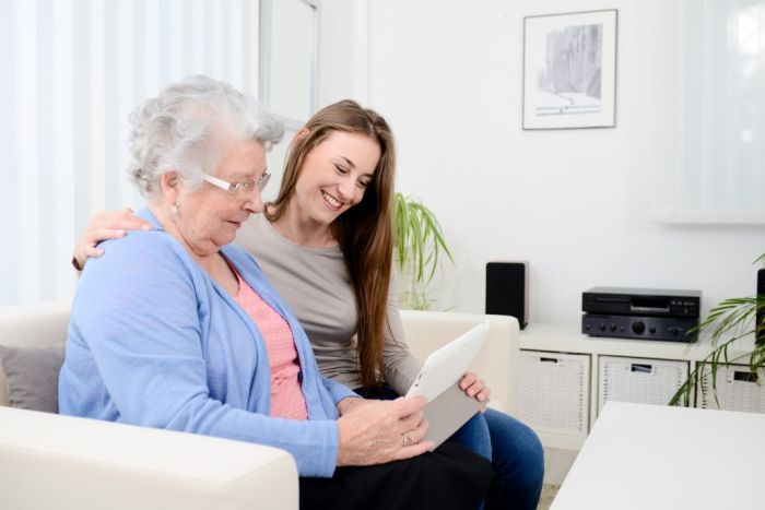 Dos mujeres están sentadas en un sofá sonriendo a una tableta en una luminosa sala de estar.