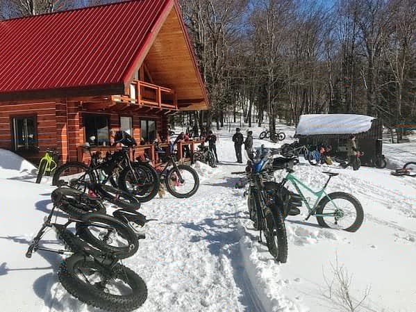 Snowy scene with fat-tire bikes parked outside a wooden cabin with a red roof. People stand near the bikes.