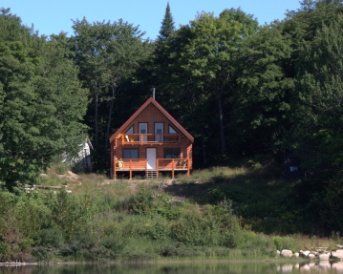 Wooden cabin on a grassy hill overlooking a body of water, surrounded by trees.