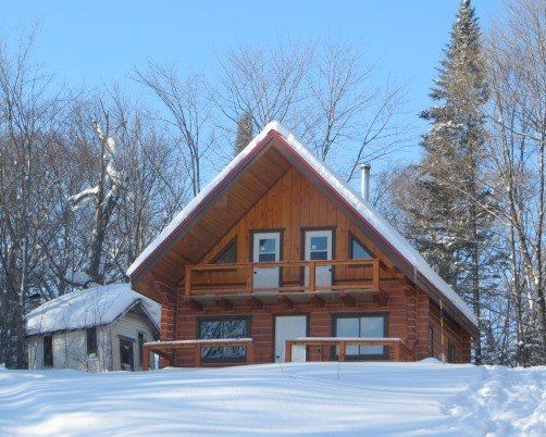 Snow-covered log cabin with a small outbuilding in a snowy forest. Blue sky, brown wood, and white snow.
