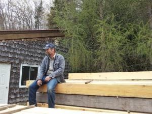 Man sits on lumber stack next to a building with a shingle roof and trees.