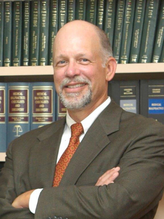 Man in a suit smiles, arms crossed. Bookshelves in the background.