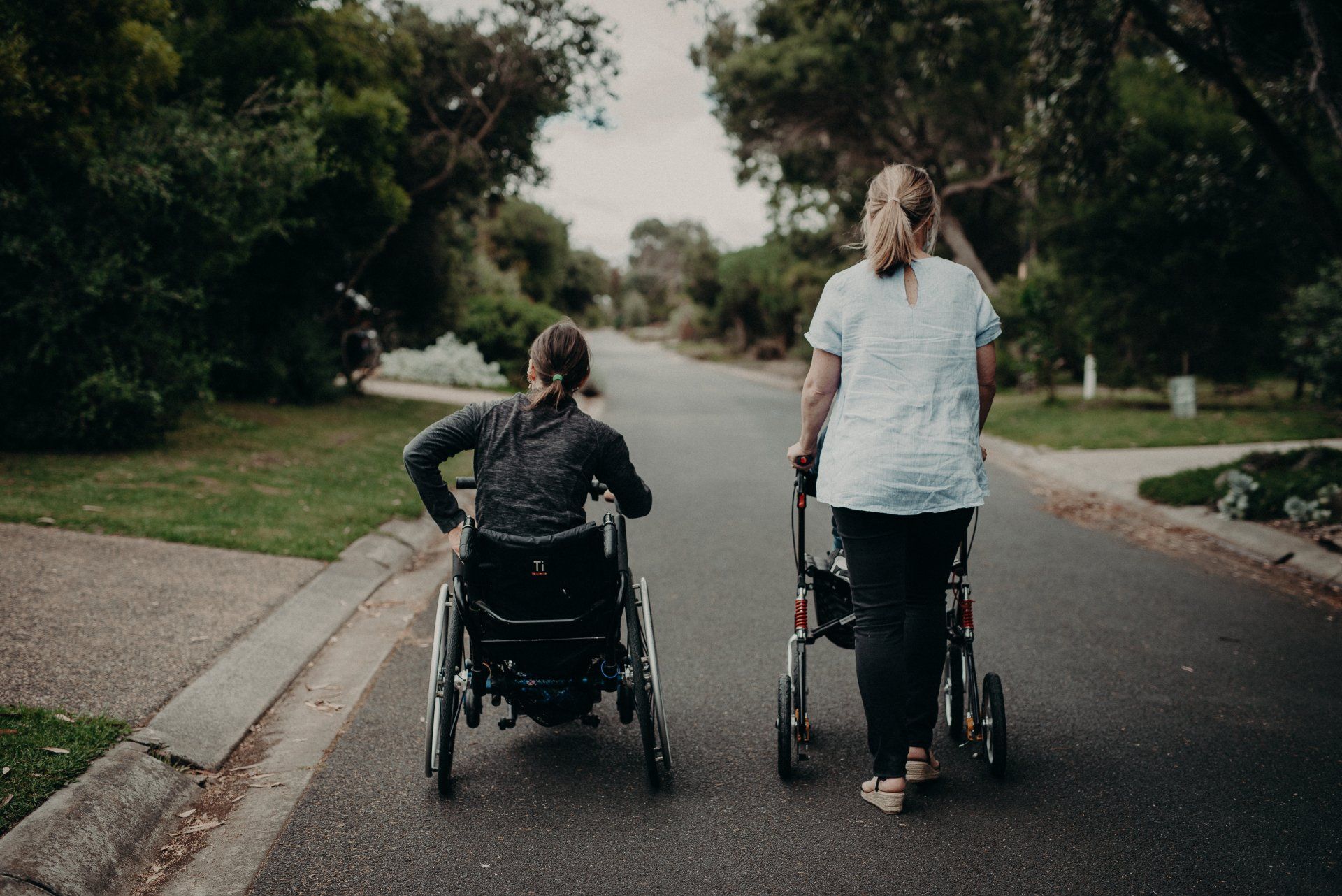 A woman in a wheelchair and a woman in a walker are walking down the street.