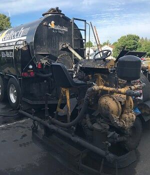 Black tanker truck with yellow and black equipment attached in a parking lot.