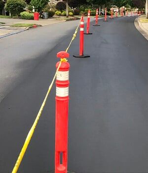 Freshly paved road, lined with orange and white traffic pylons and yellow caution tape.