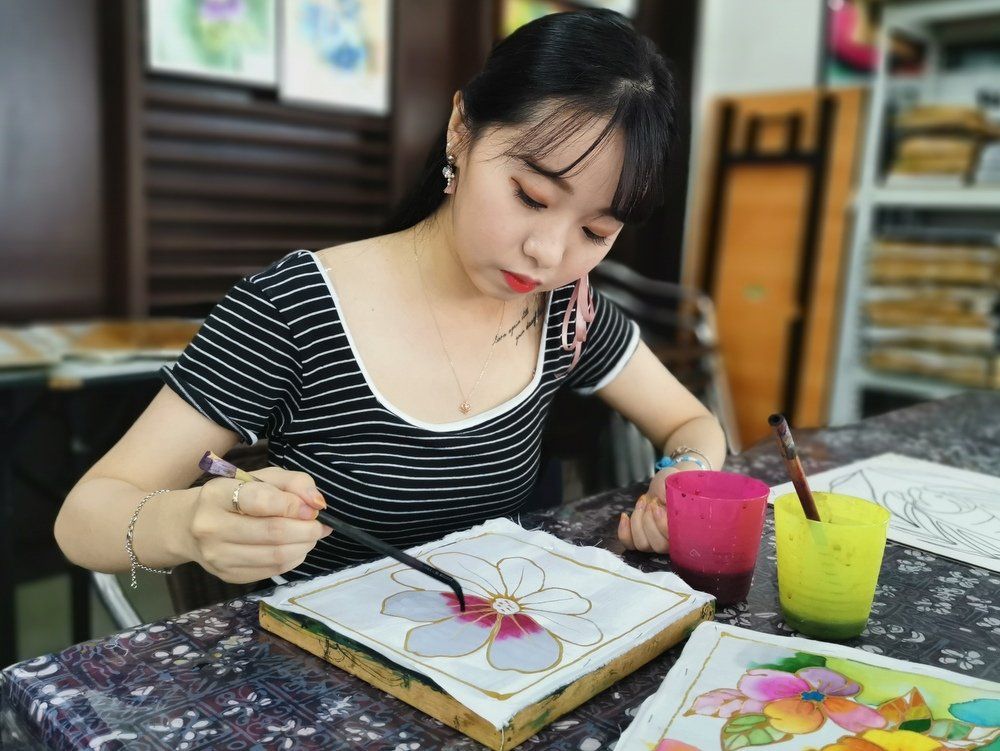 Woman painting a flower design, seated at a table. She wears a striped shirt, holding a brush with focused expression.