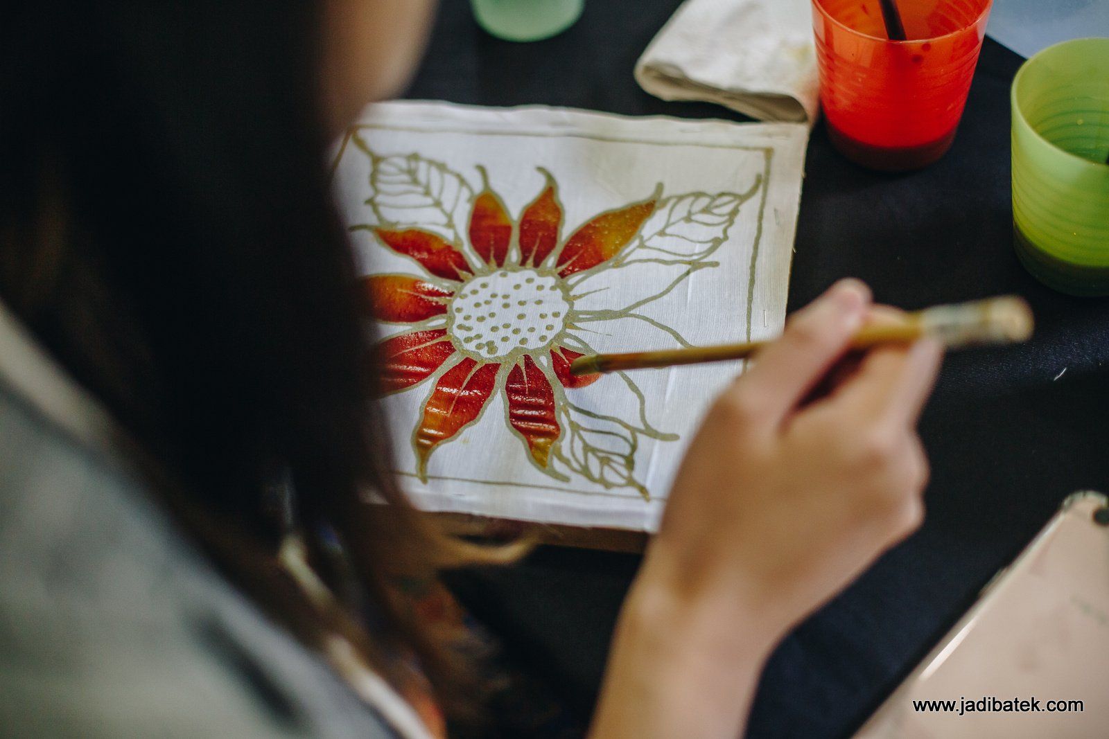 Person painting a flower design on fabric with orange and red paint, a paintbrush and a cup of liquid nearby.