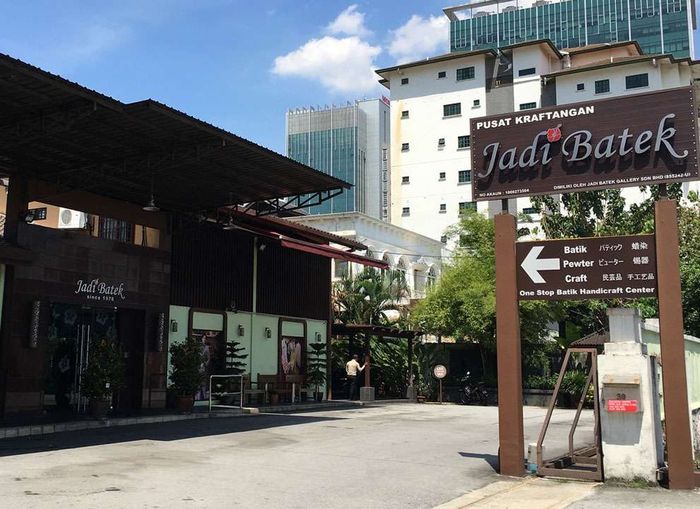 Sign for Jadi Batek, a batik shop, pointing left. Buildings and a blue sky in the background.
