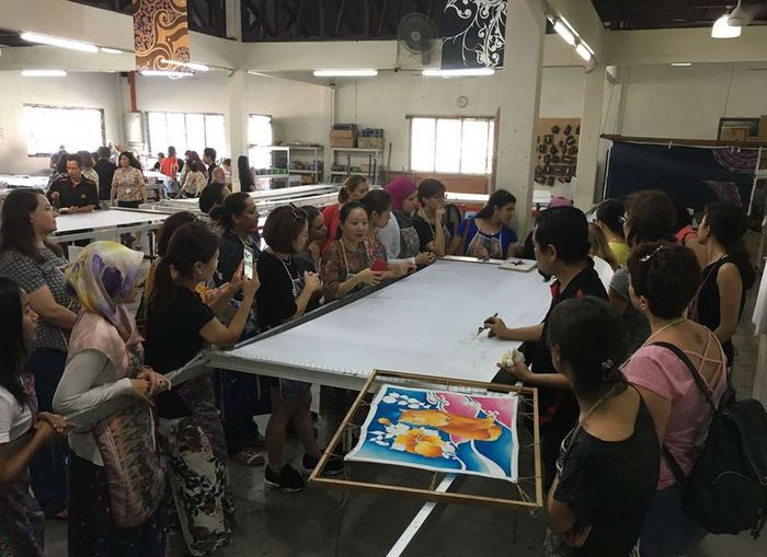 People observing a batik artist drawing on a large white canvas in a workshop.