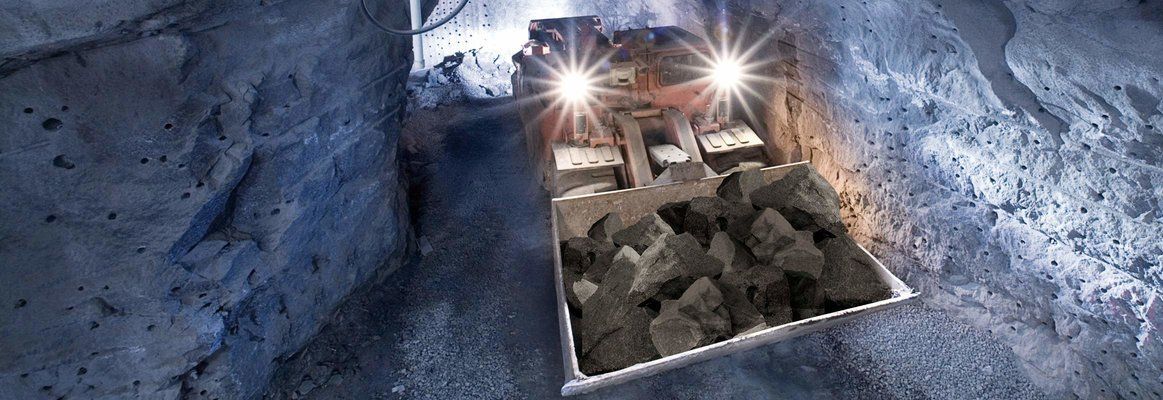 A mining loader machine with bright headlights hauls a scoop of gray rock through a dark, narrow underground tunnel.
