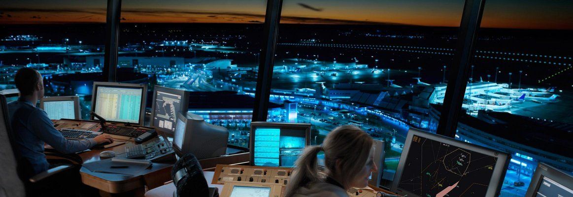 Two air traffic controllers work at monitors in a dark control tower overlooking a lit airport runway at night.