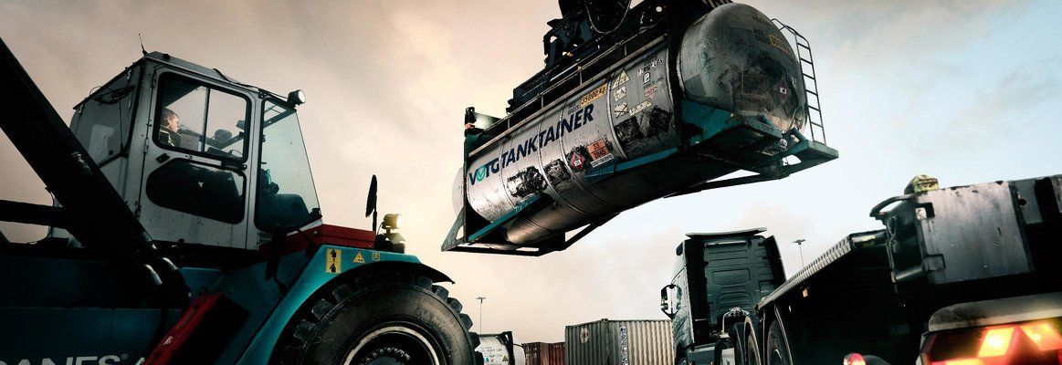 A reach stacker lifts a large, cylindrical cargo container at a busy shipping port during twilight.