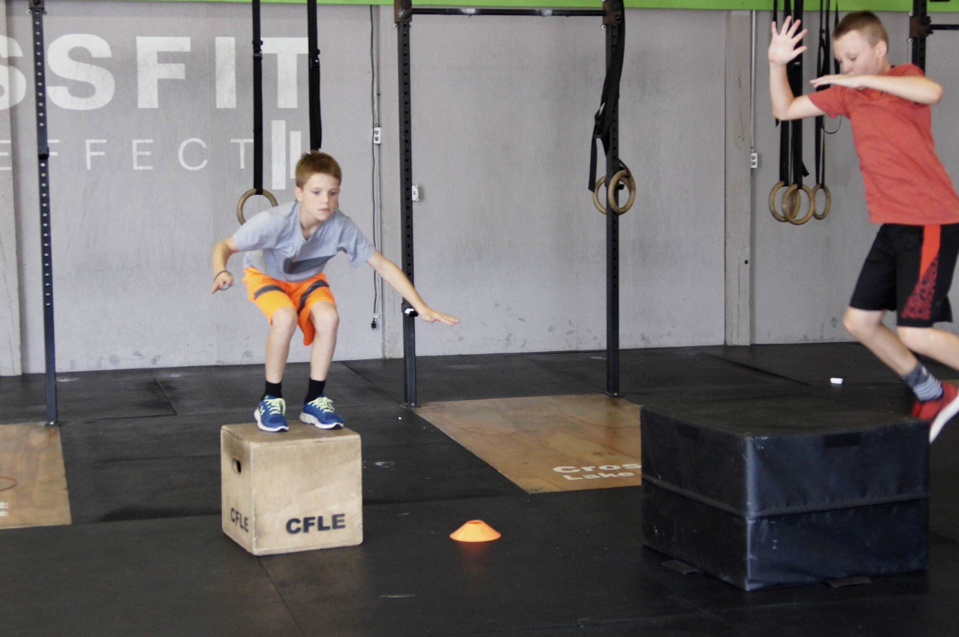 Two young boys are jumping over boxes in a gym with the word crossfit in the background