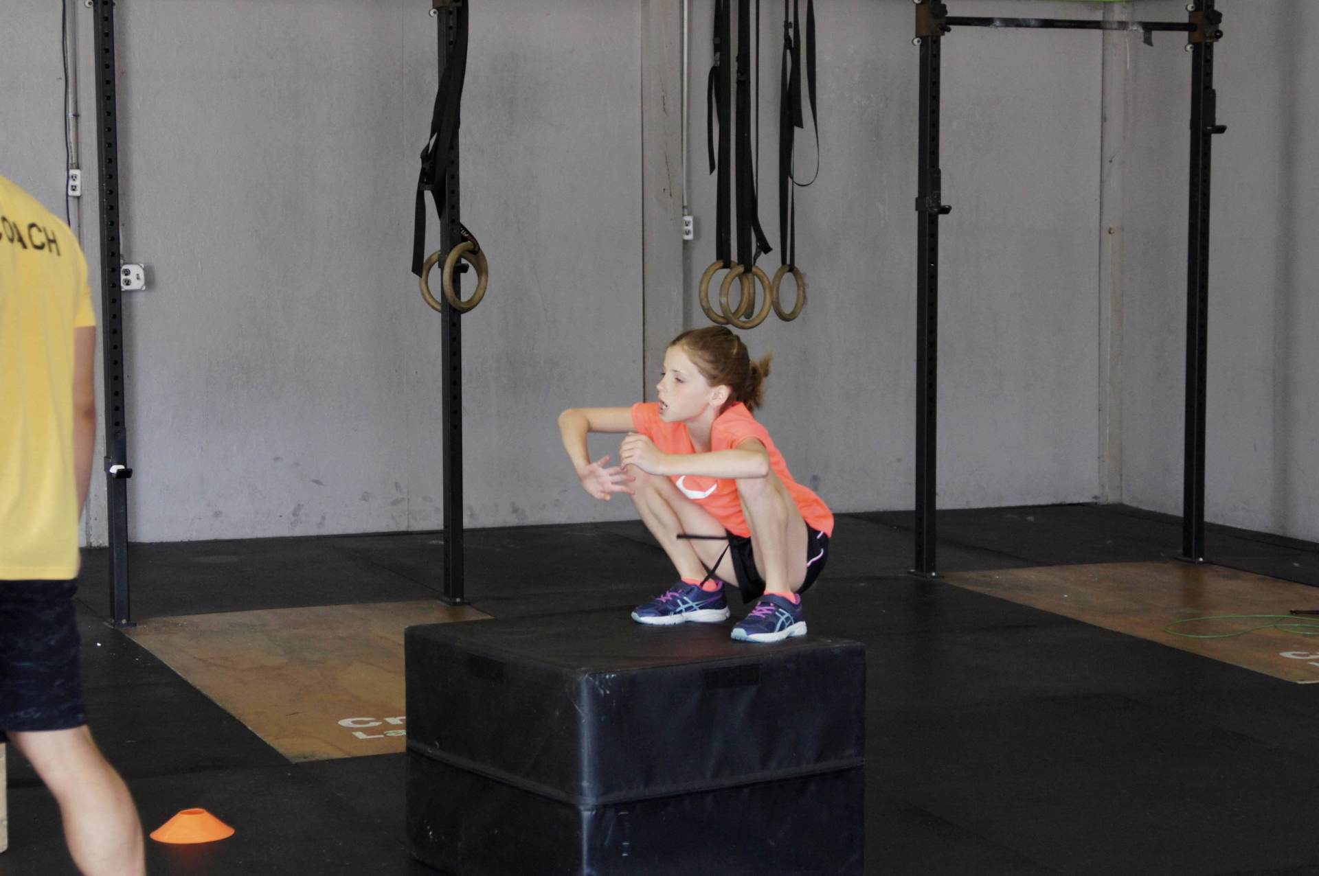A little girl is squatting on a box in a gym.