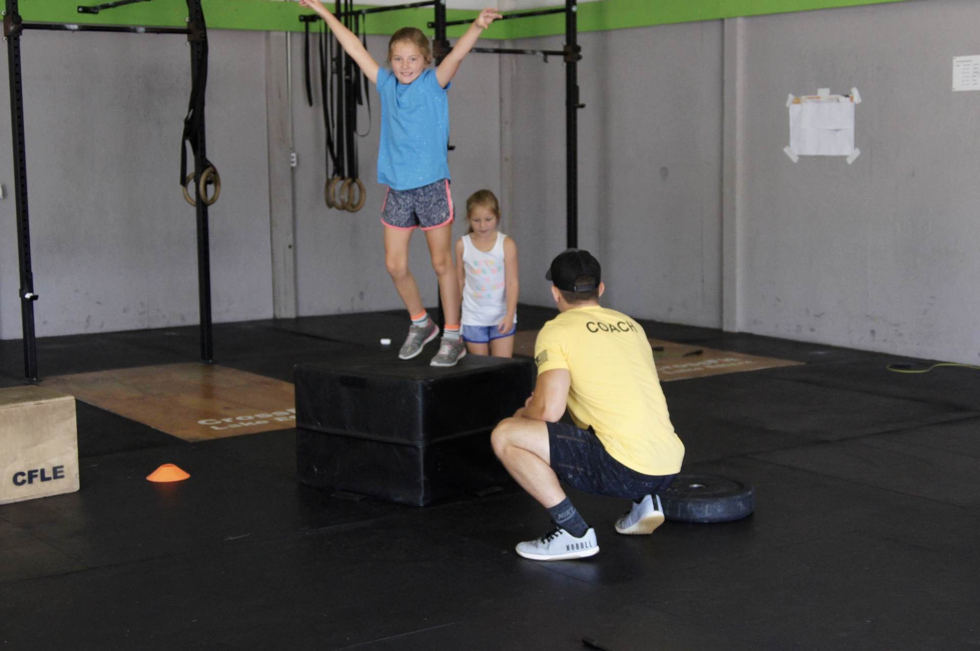 A man in a yellow shirt is kneeling next to a girl standing on a box in a gym.