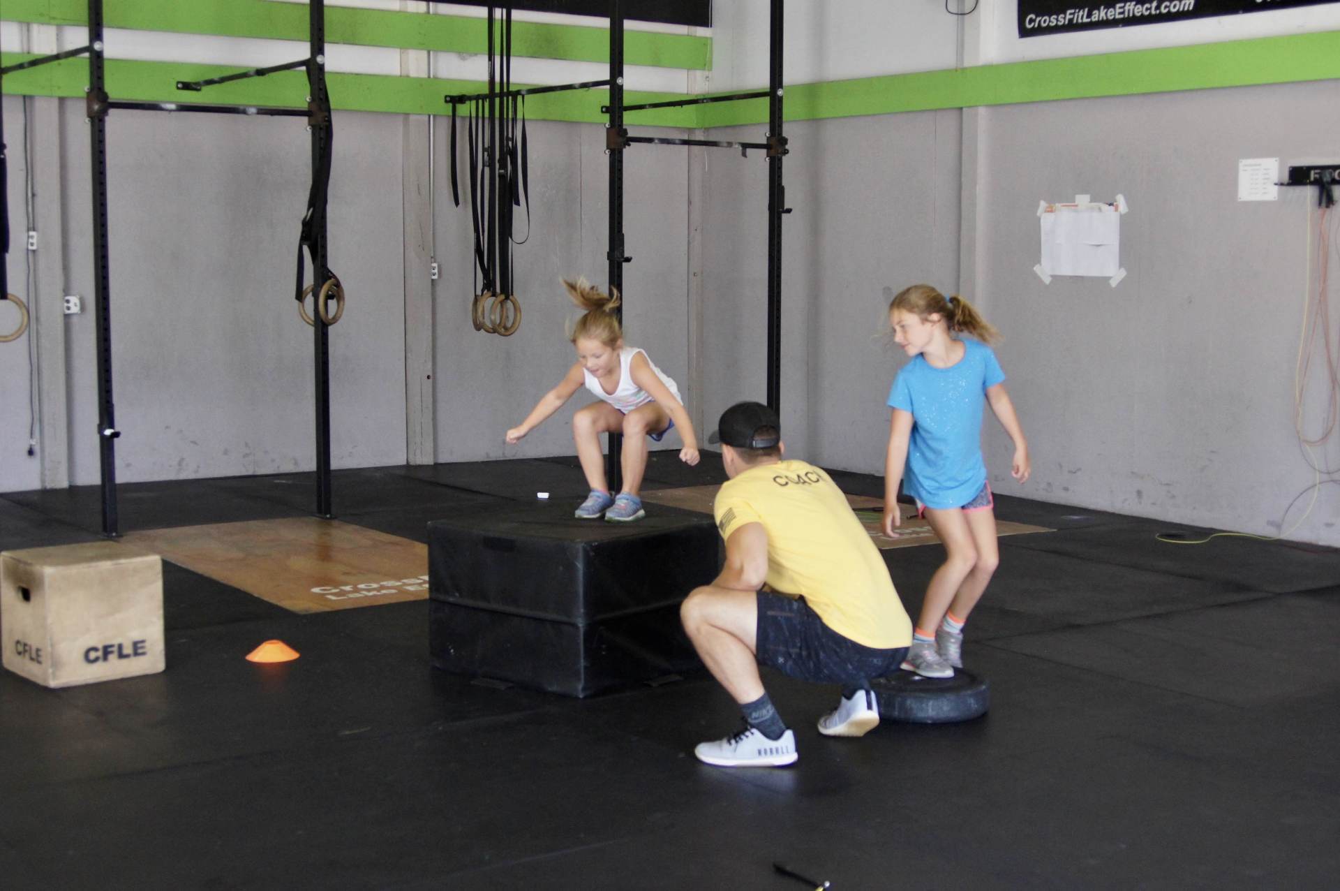 A man is squatting down next to two girls who are jumping on boxes in a gym.