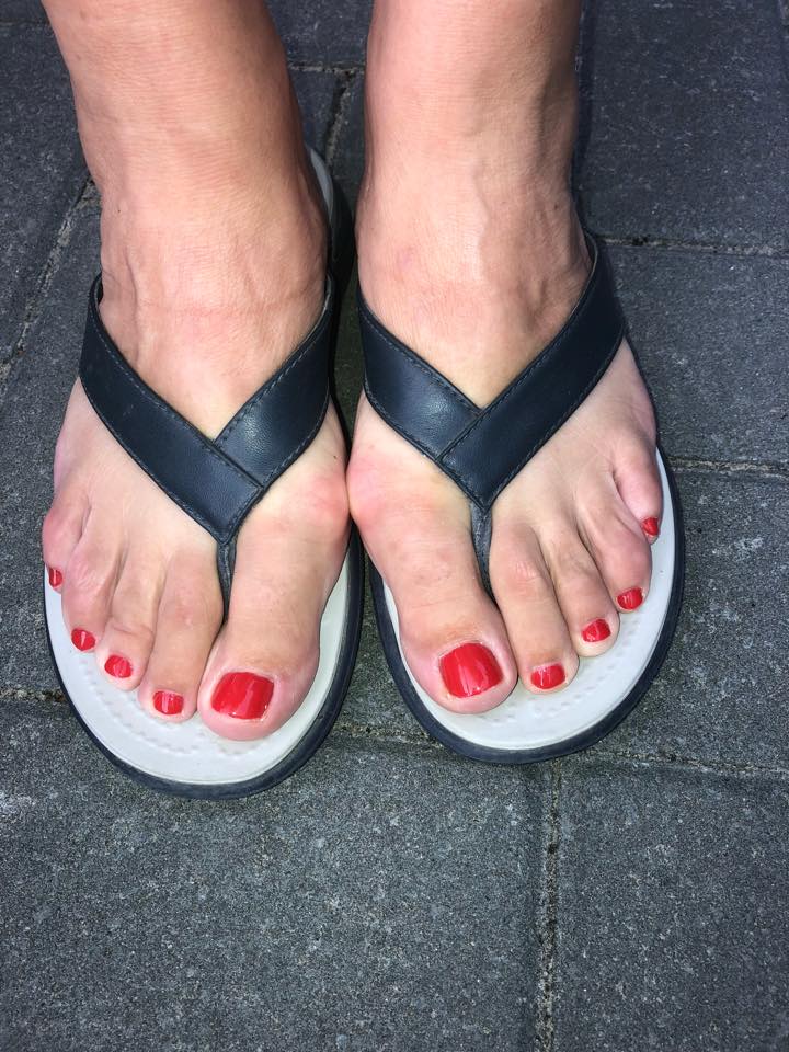 Voeten in zwarte teenslippers met rode nagellak op de tenen staan ​​op een donker trottoir.