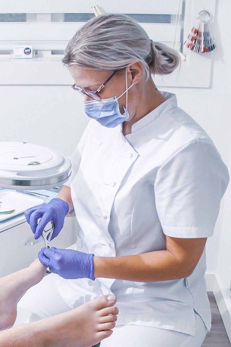 Een zorgmedewerker in een wit uniform en paarse handschoenen behandelt de voet van een patiënt in een kliniek.