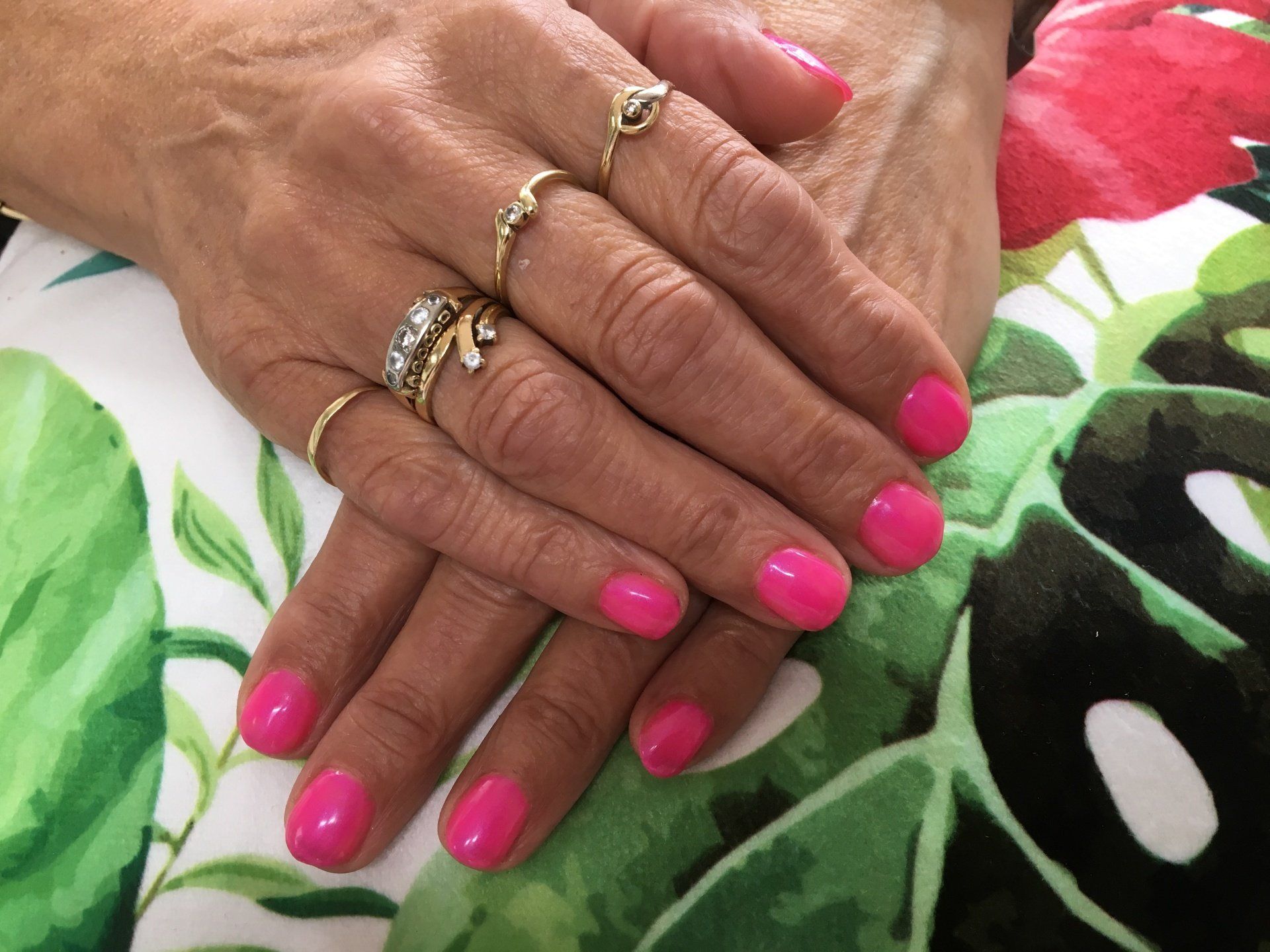 Handen met felroze nagels en gouden ringen rusten op een stof met bloemenpatroon.