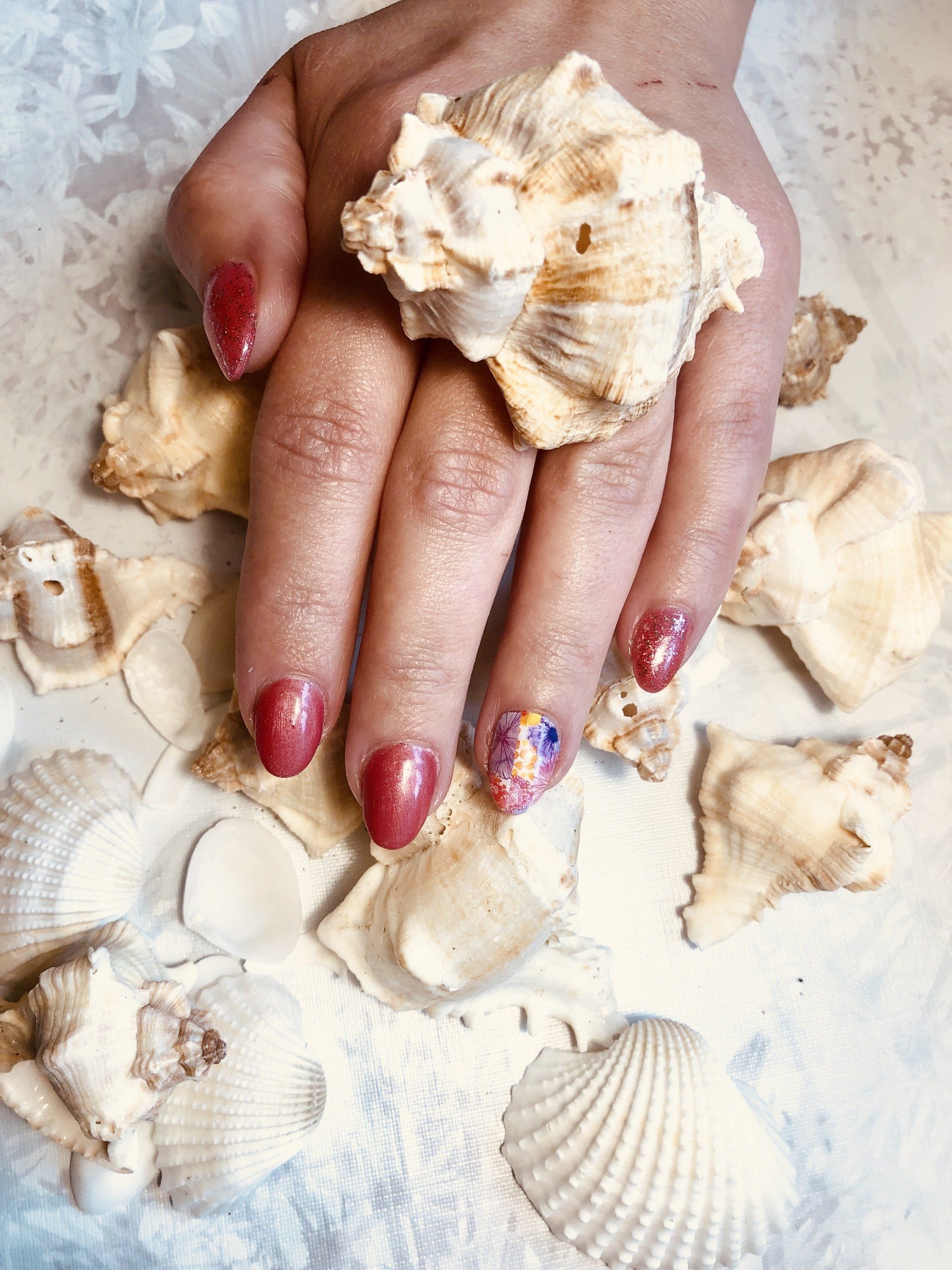 Hand met rode en glinsterende nagels rustend op schelpen en zand.