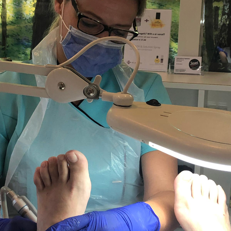 Een podoloog met blauwe handschoenen en een mondkapje onderzoekt de voeten van een patiënt onder een lamp.