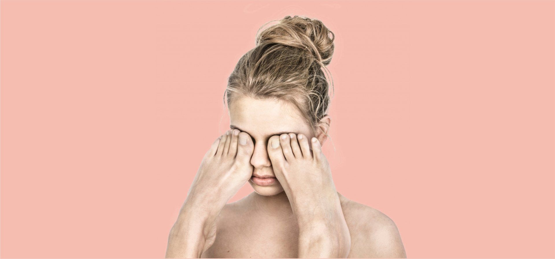 Vrouw met opgestoken haar wrijft in haar ogen tegen een roze achtergrond.