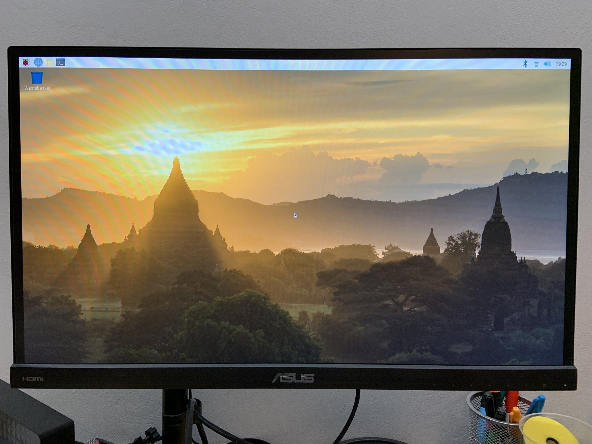 An ASUS computer monitor displaying the Raspberry Pi desktop OS with a sunset landscape featuring ancient temples.