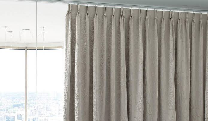 Beige pleated drapes hang from a curtain rod in front of a window.