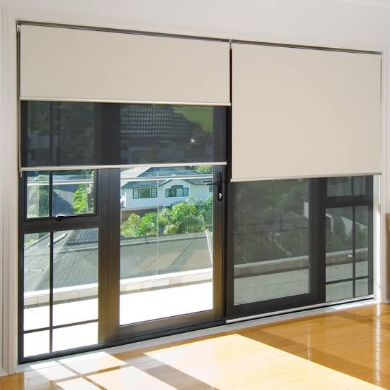 Two roller blinds over a glass door and window, one lowered, one raised, in a room with wood flooring.