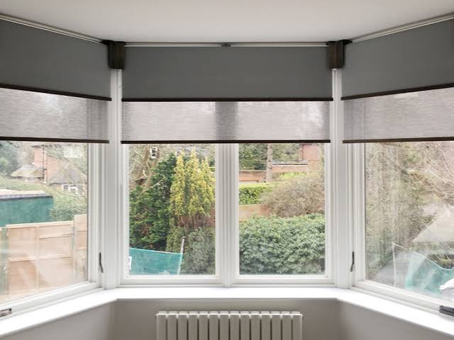 Gray roller blinds cover a bay window, looking out onto a garden with trees and a radiator below.