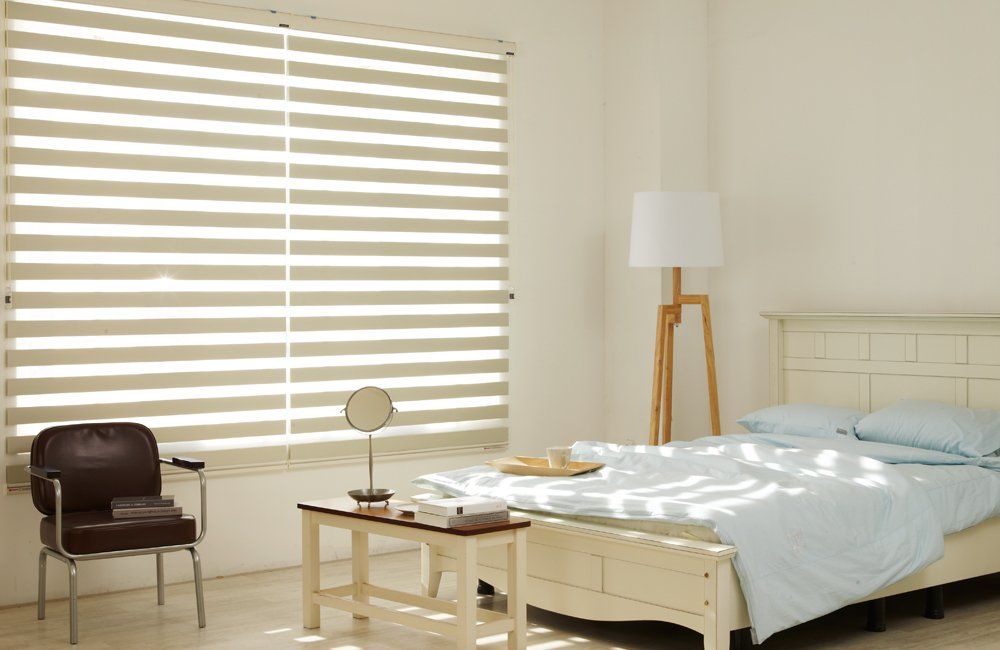 Bedroom with a bed, a window with blinds, a chair, a side table with a mirror, and a floor lamp.
