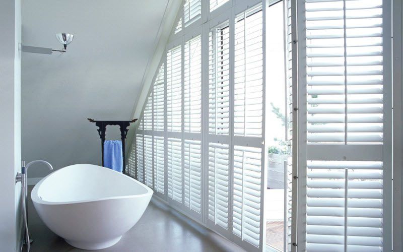 White bathroom with oval tub, angled ceiling, and shuttered windows.