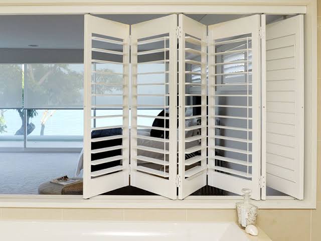 White folding window shutters in a room with a view, partly open, beige surfaces.