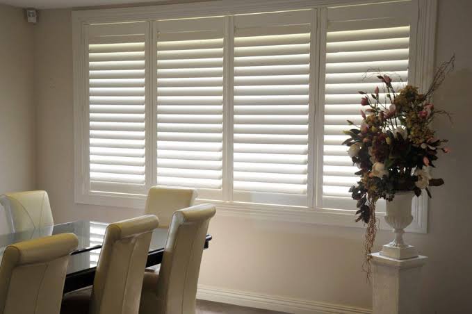 White shuttered windows in a dining room, with a dark table, cream chairs, and floral arrangement.