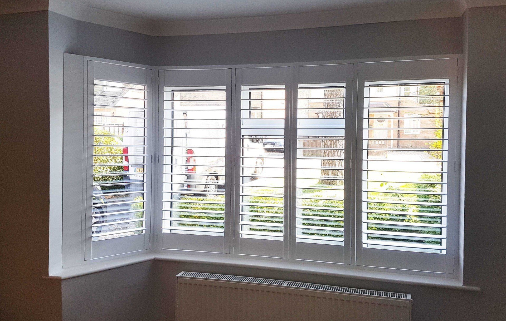 White shutters cover a bay window, with a radiator below. The window looks out to a blurry outdoor scene.