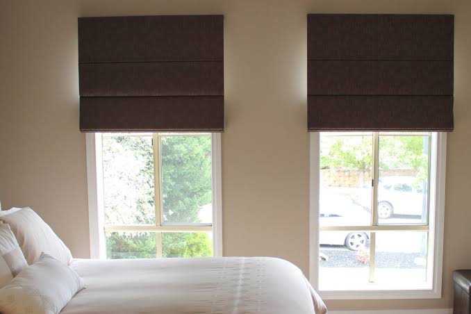 Bedroom with bed, two windows with brown Roman shades. Outside view of trees and car.