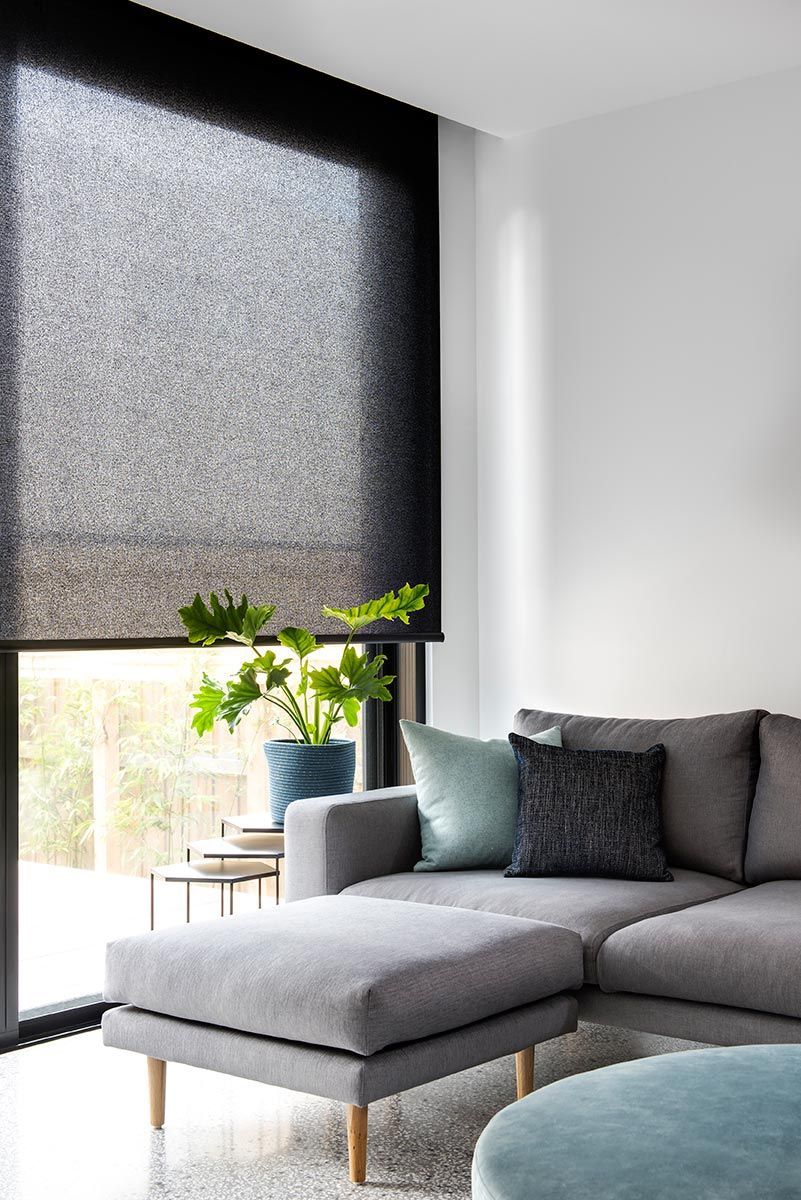 Living room with grey couch, ottoman, and dark roller blind. A potted plant sits near the window.