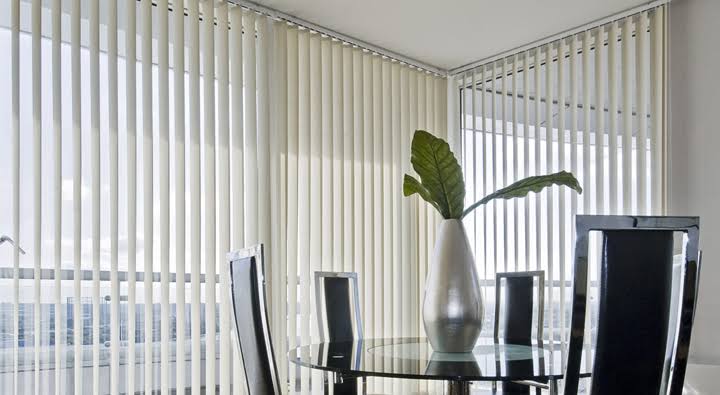 Dining room with vertical blinds, glass table, black chairs, and a vase with green leaves.