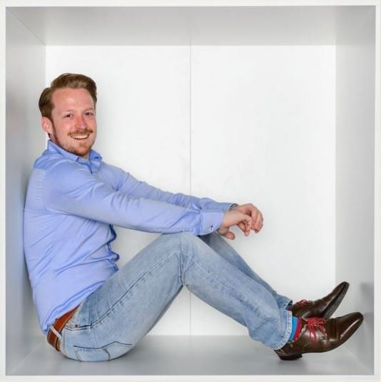 Een glimlachend persoon in een blauw overhemd en spijkerbroek zit in een spierwitte, vierkante ruimte.