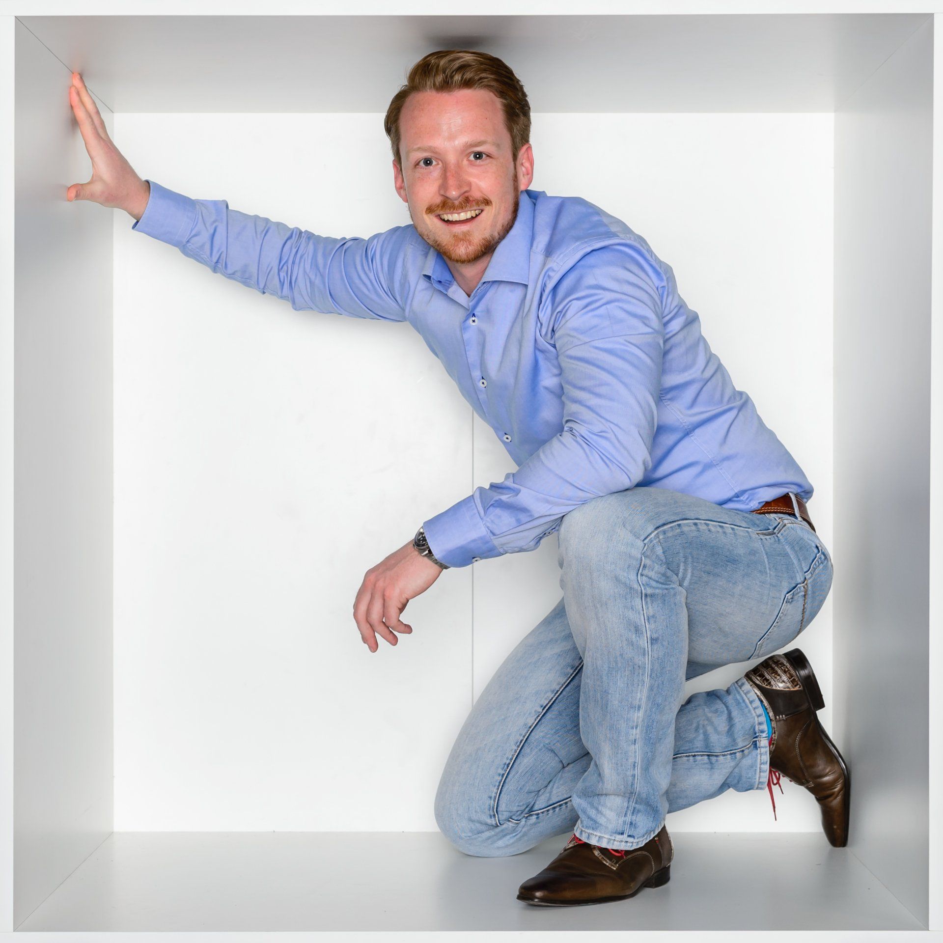 Een glimlachend persoon in een blauw shirt en spijkerbroek hurkt in een wit, vierkant kantoorhokje en raakt met één hand de zijwand aan.