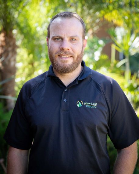 Man with a beard in a black polo shirt smiles outside. Green foliage in the background.