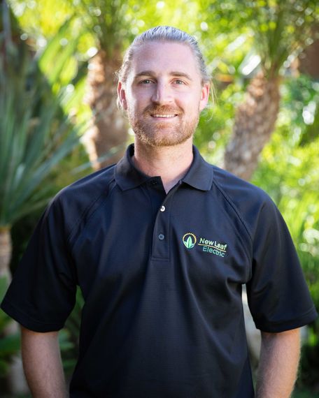 Man with light brown hair in a black polo shirt smiles outside with palm trees in the background.